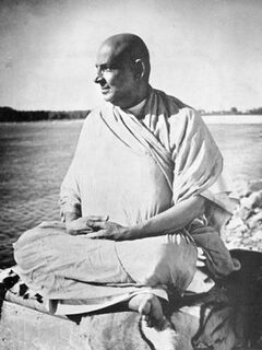 Swami Sivananda sitzt mit gekreuzten Beinen am Wasser